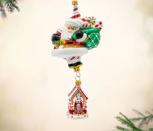Santa Claus ornament carrying a green gift sack above a small gingerbread house.