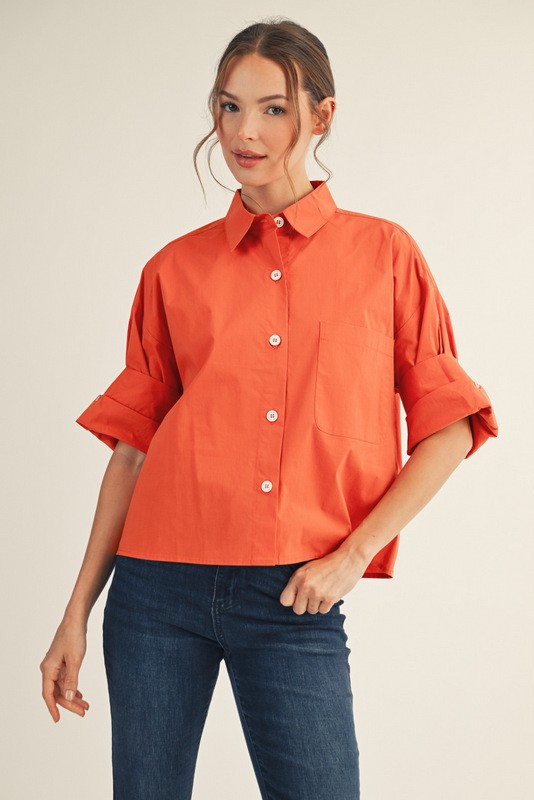 Woman wearing an orange Clarisse multi-fold sleeve button-down shirt with dark blue jeans.