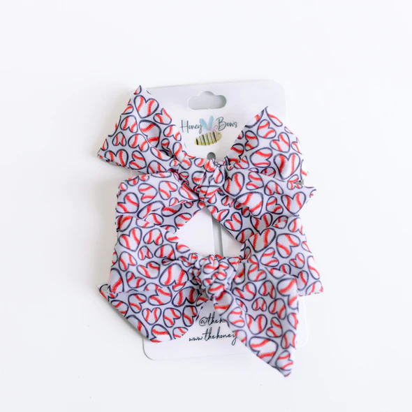 Two 3.5-inch hair bows on a white card featuring a heart-shaped baseball pattern with red stitching.