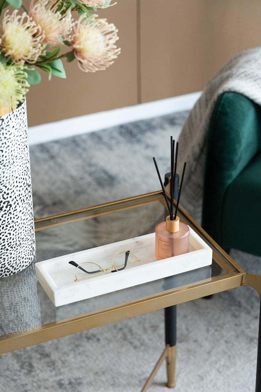 Rectangular white marble tray on a glass table holding gold glasses and a reed diffuser.