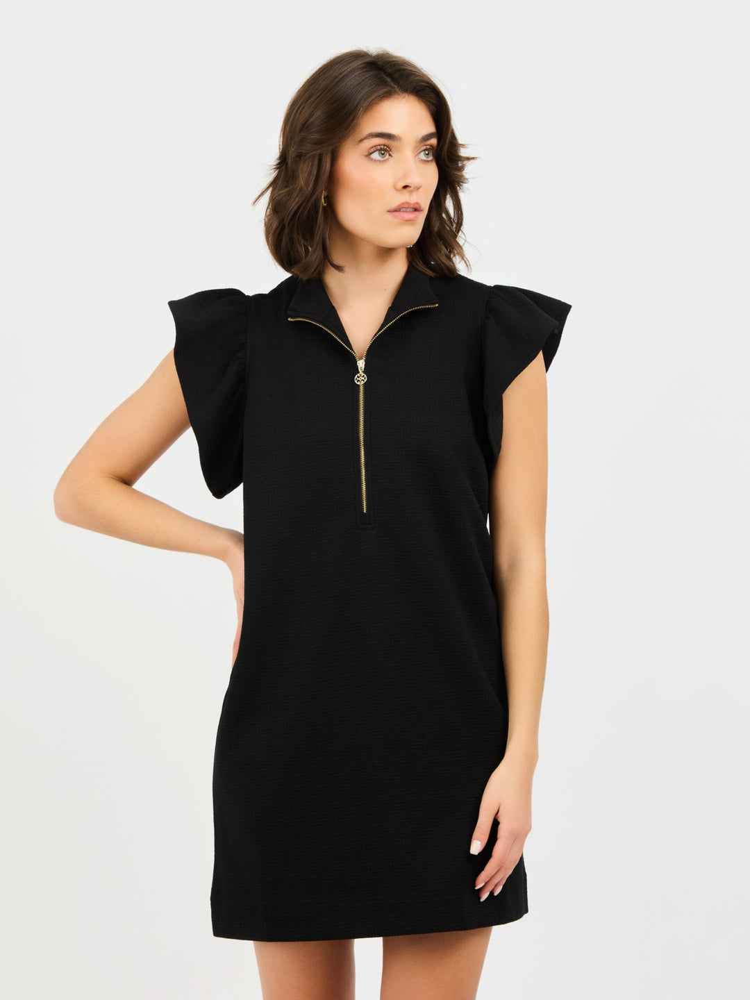 Woman wearing the Paige Dress, a black mini dress with flutter sleeves and a gold front zipper.