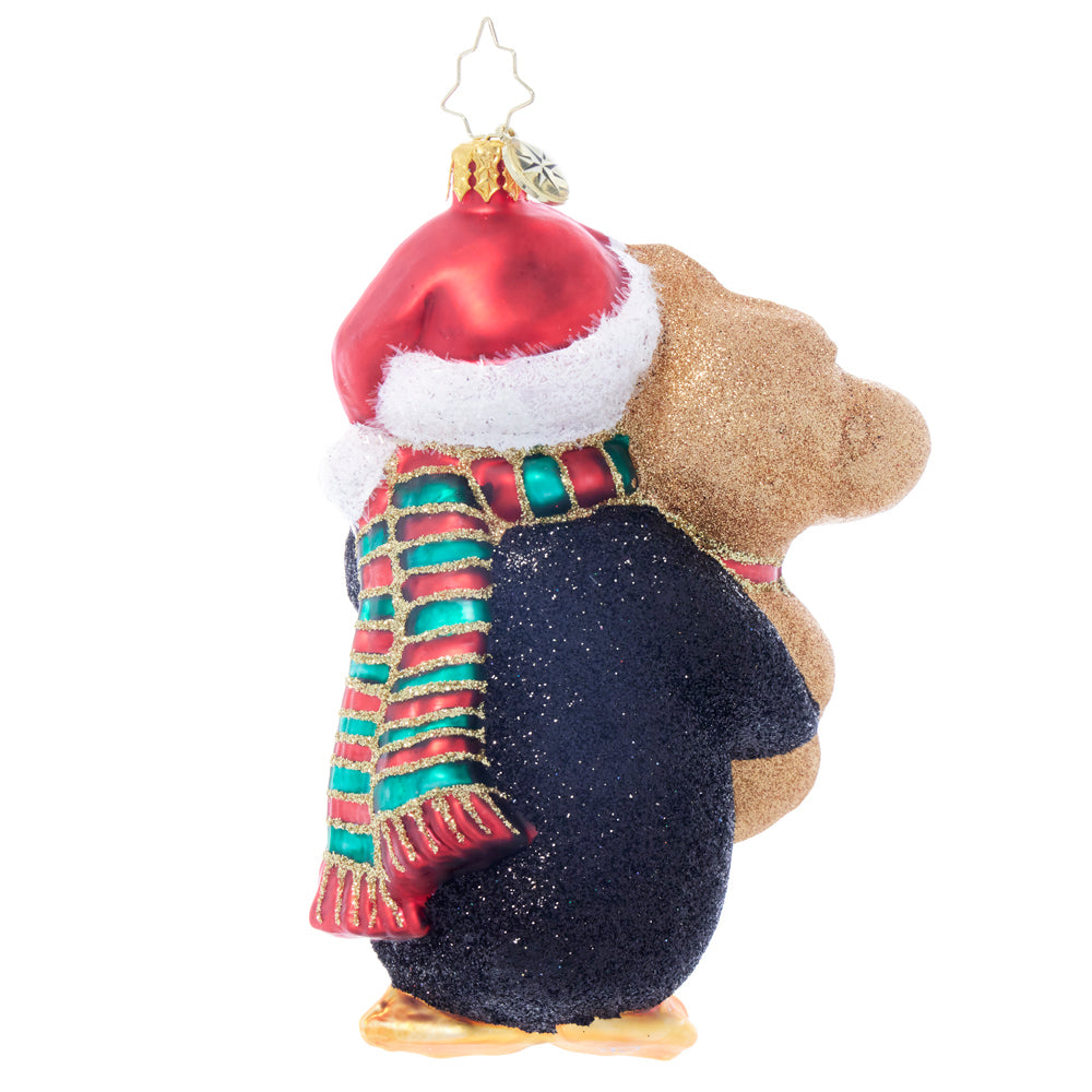 Side view of a penguin Christmas ornament wearing a Santa hat and scarf, hugging a teddy bear.