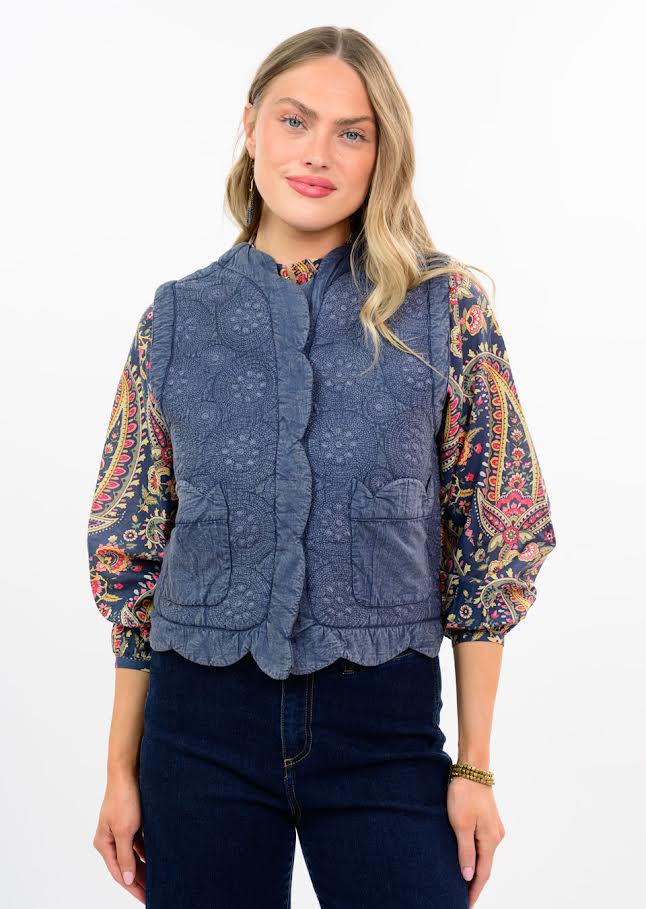 Woman wearing the Amelia blue embroidered scalloped vest over a paisley print long-sleeve top.