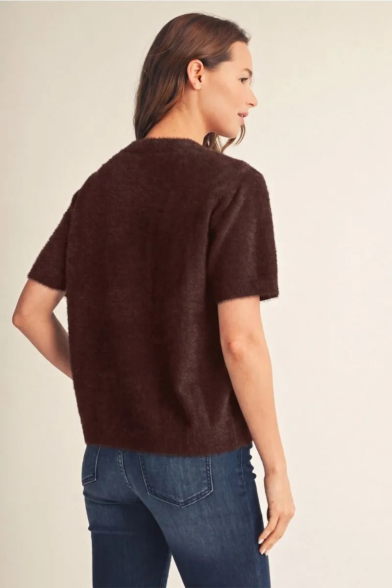 Back view of a woman wearing a dark brown, short-sleeved fuzzy sweater cardigan with blue jeans.