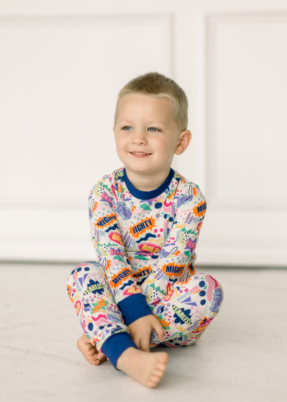 Young boy sitting in The Mighty Mack bamboo pajamas with colorful comic-style words and patterns.