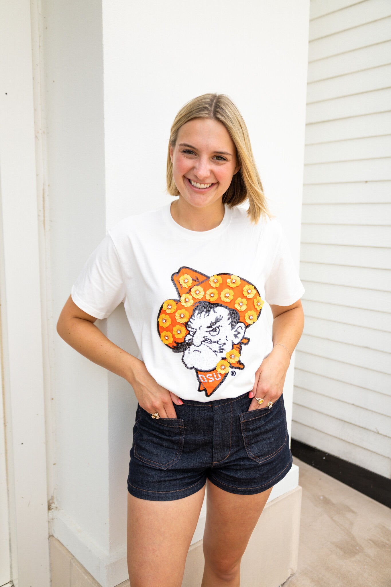 Woman in a white Arah Licensed Oklahoma State Mascot Sequin Flower Tee and dark denim shorts.
