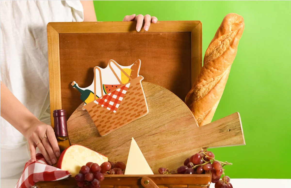 Brown ceramic picnic basket attachment with red buffalo print napkin holding wine, cheese, fruit and bread.