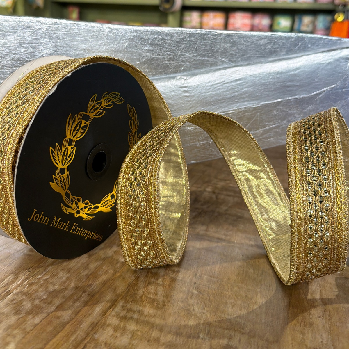 1x10yd Jeweled Trim Metallic Dupion Gold Ribbon on a black spool with a gold laurel wreath logo.