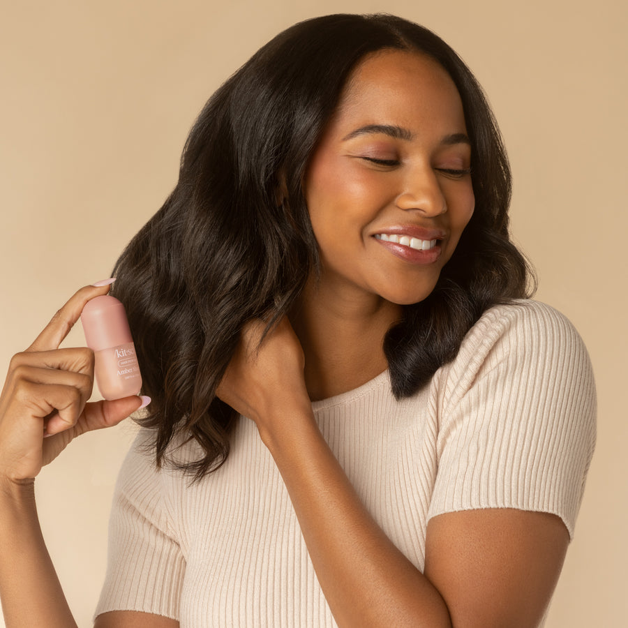 A woman smiling and holding a Mini Amber Shores Hair & Body Perfume Mist bottle near her hair.