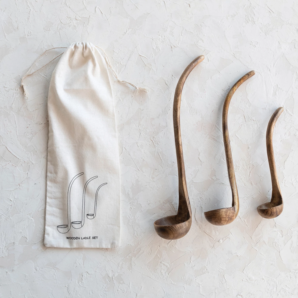 Three mango wood ladles in varying sizes with a walnut finish next to a printed drawstring bag.