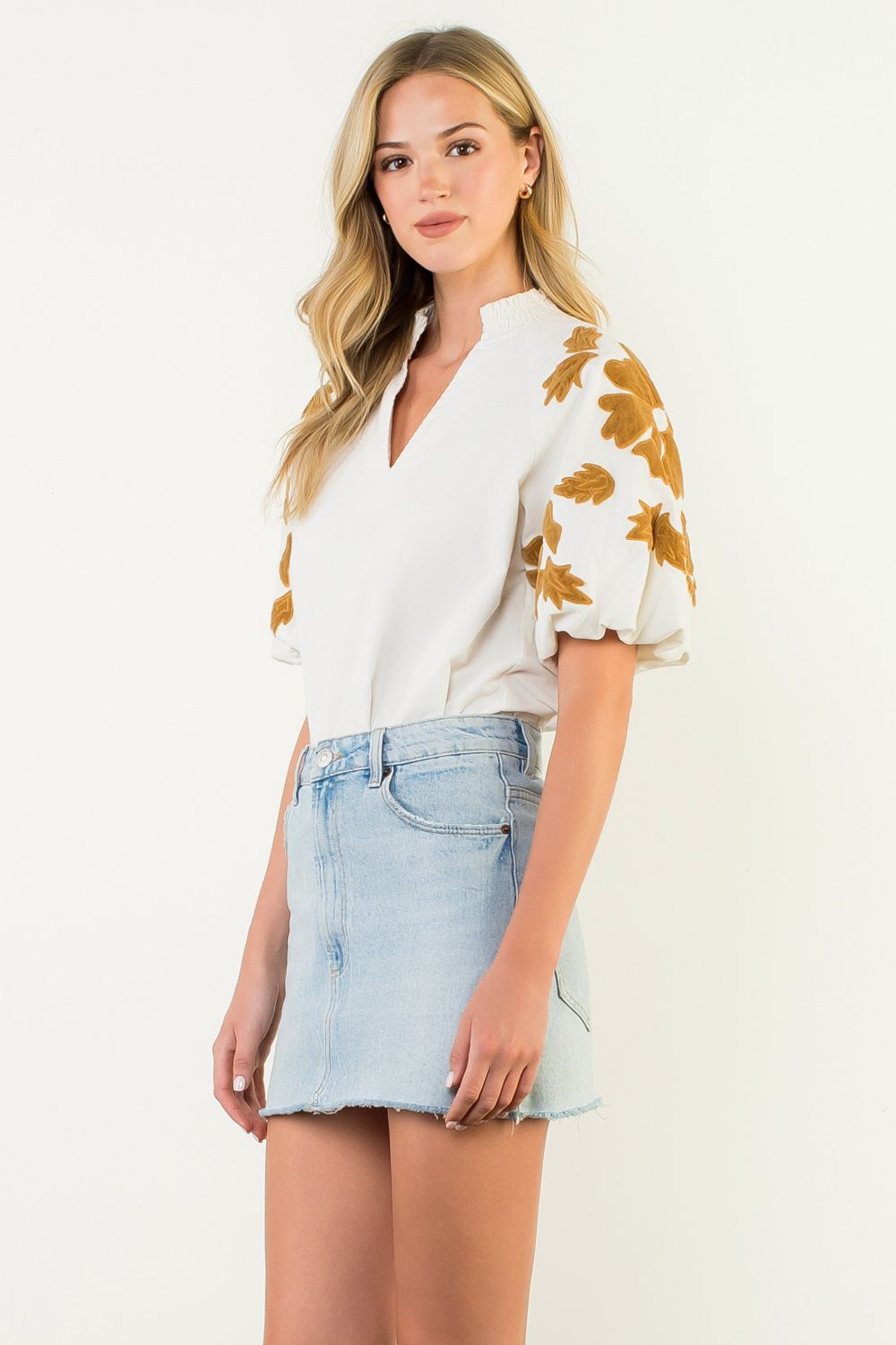 Woman wearing a white Sophia top with gold chenille floral puff sleeves and a denim mini skirt.