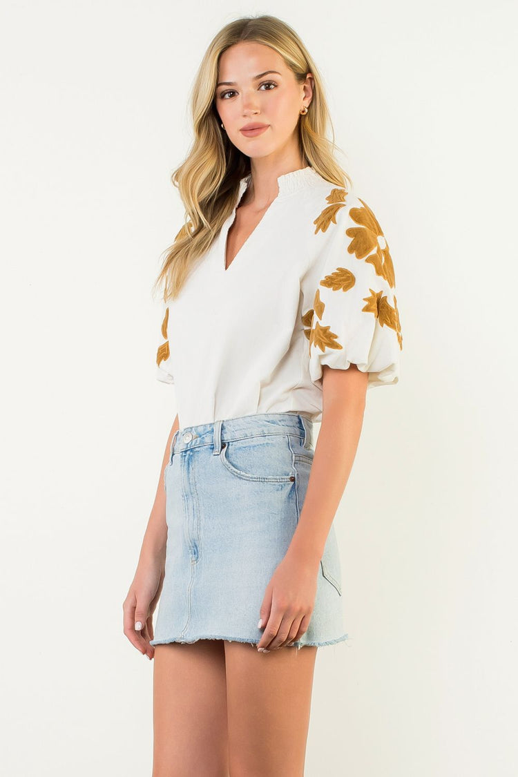 Woman wearing a white Sophia top with gold chenille floral puff sleeves and a denim mini skirt.