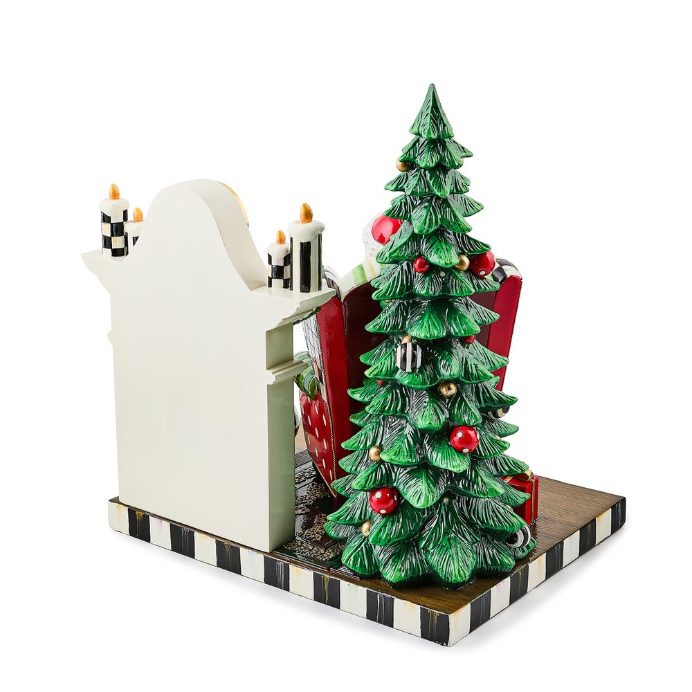 Rear view of a Santa figurine by a fireplace with a decorated Christmas tree on a striped base.
