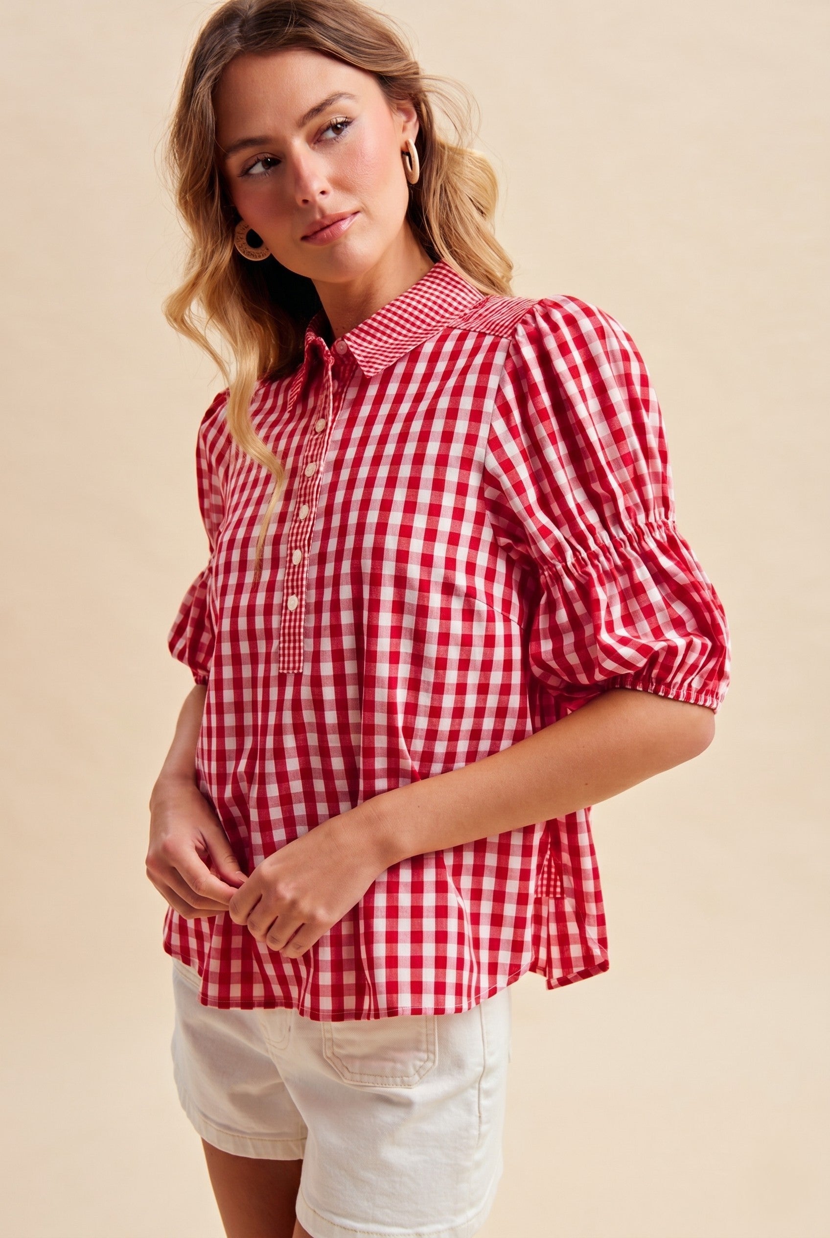 Woman wearing a red and white Suzy Gameday Gingham Puff Sleeve Henley Blouse with white shorts.