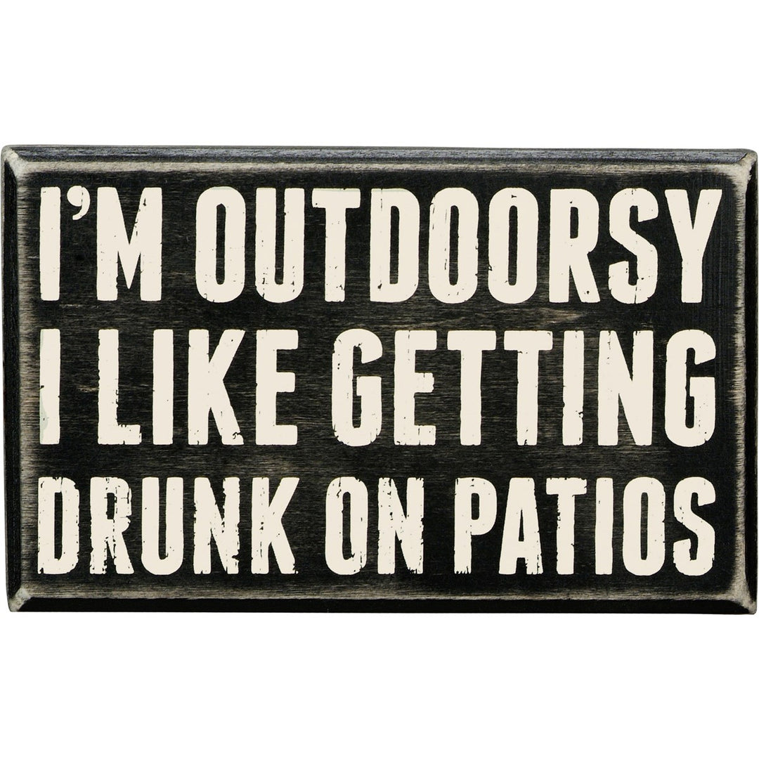 Black wooden box sign with white text that reads "I'M OUTDOORSY I LIKE GETTING DRUNK ON PATIOS."