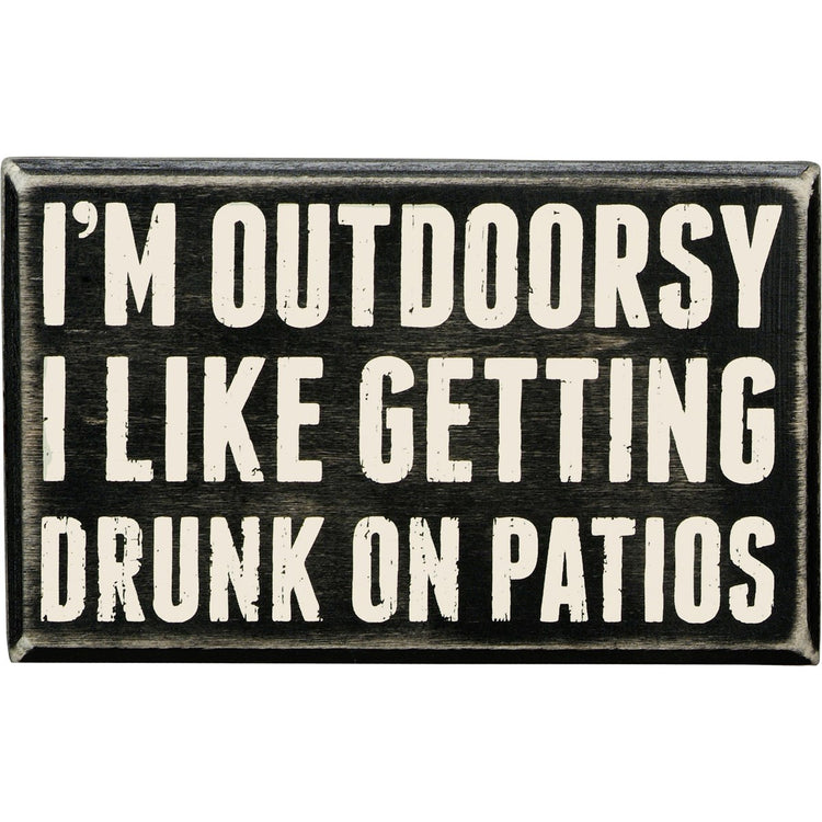Black wooden box sign with white text that reads "I'M OUTDOORSY I LIKE GETTING DRUNK ON PATIOS."