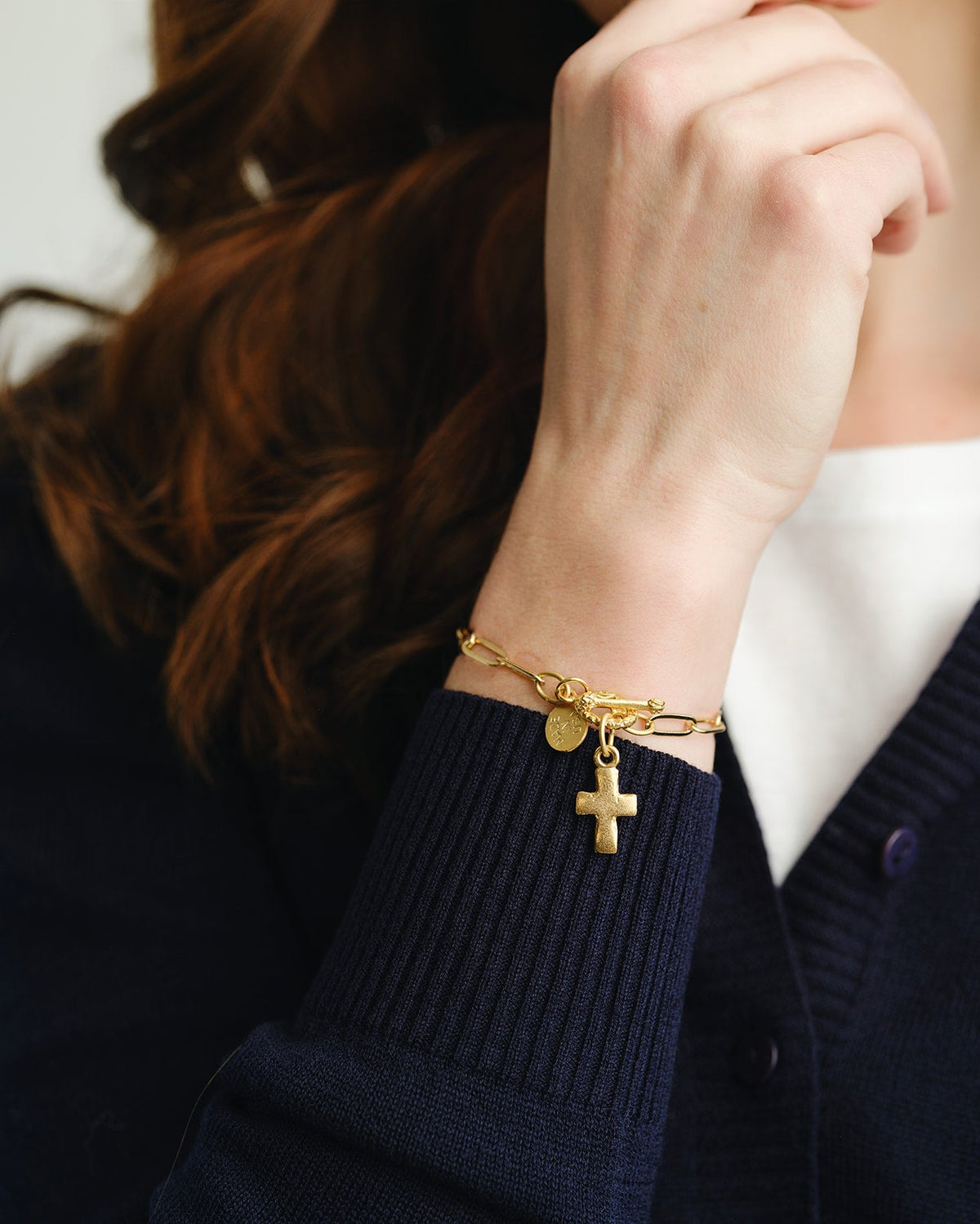 A person wearing a gold paperclip chain bracelet with a small cross pendant and toggle clasp.