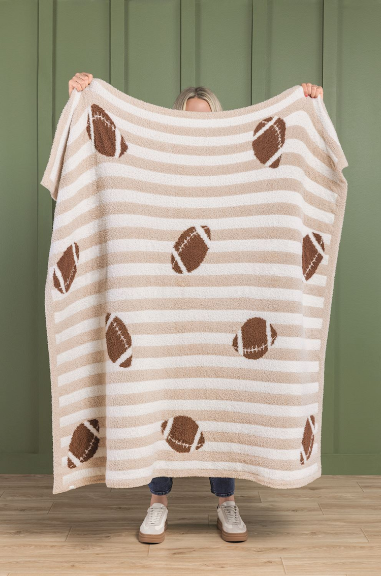 Person holding a tan and white striped chenille blanket with a brown football pattern.