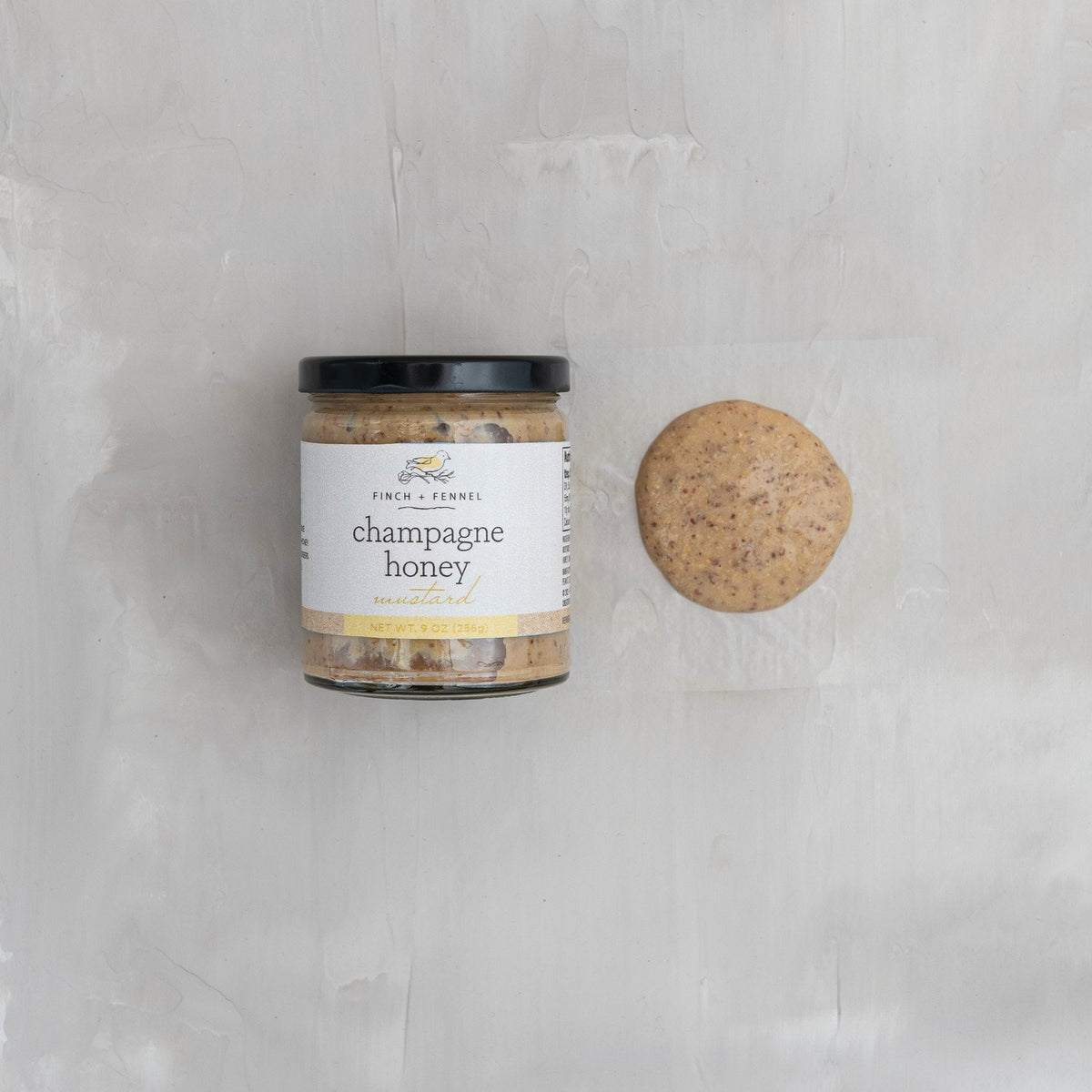 A jar of Finch + Fennel Champagne Honey Mustard next to a dollop of the grainy mustard.