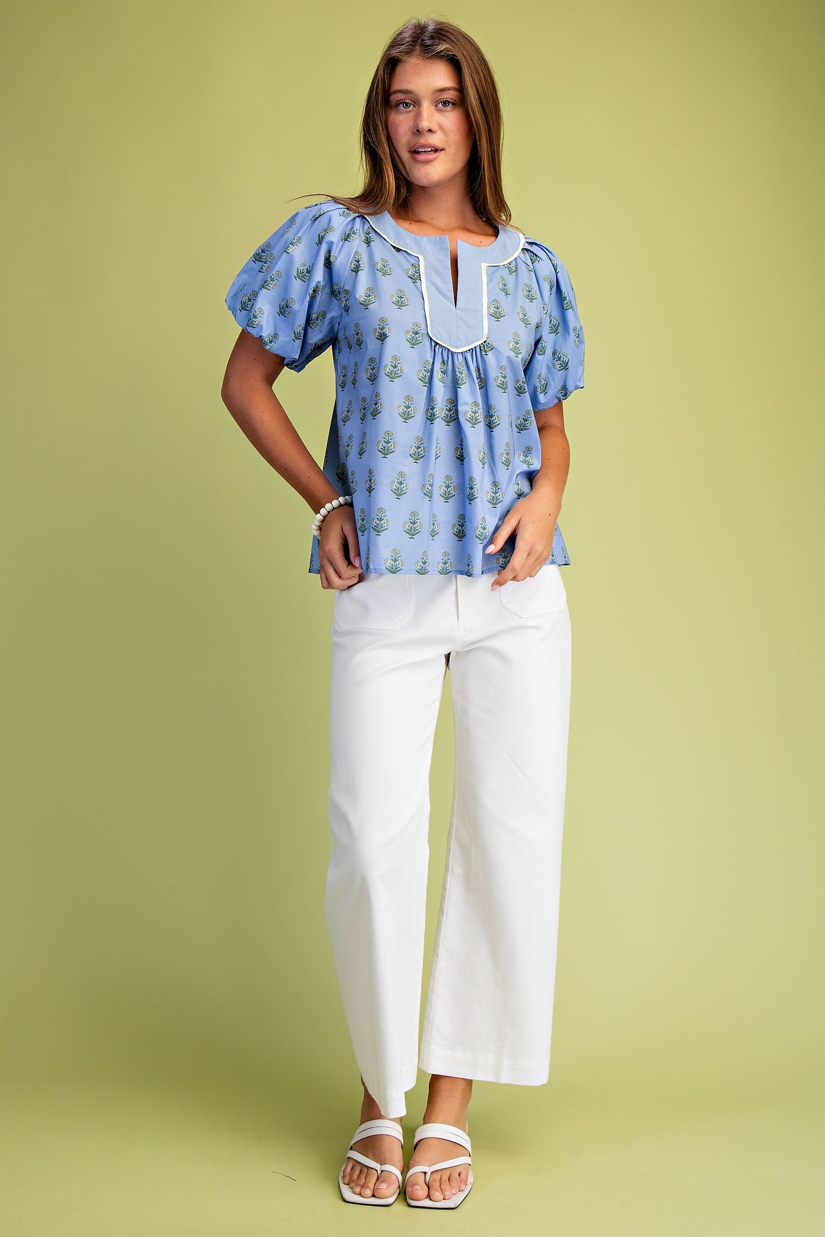 Woman wearing a blue floral notch neck pleated blouse with short puff sleeves and white pants.