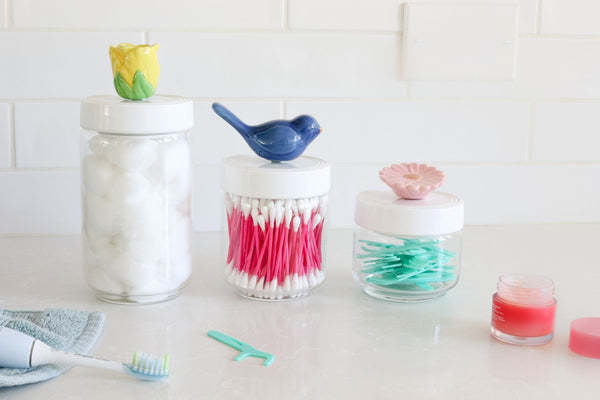 Three glass jars with decorative flower and bird lids, filled with cotton balls and dental tools.