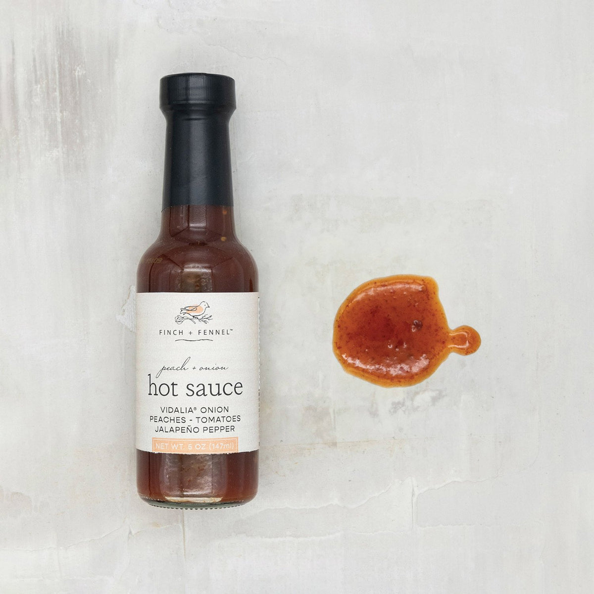 A 5oz bottle of Finch + Fennel Peach + Onion Hot Sauce next to a sample of the orange-red sauce.