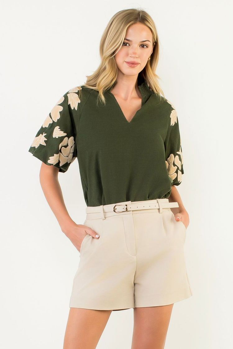 Woman wearing a Sophia olive green top with embroidered chenille floral puff sleeves and tan shorts.