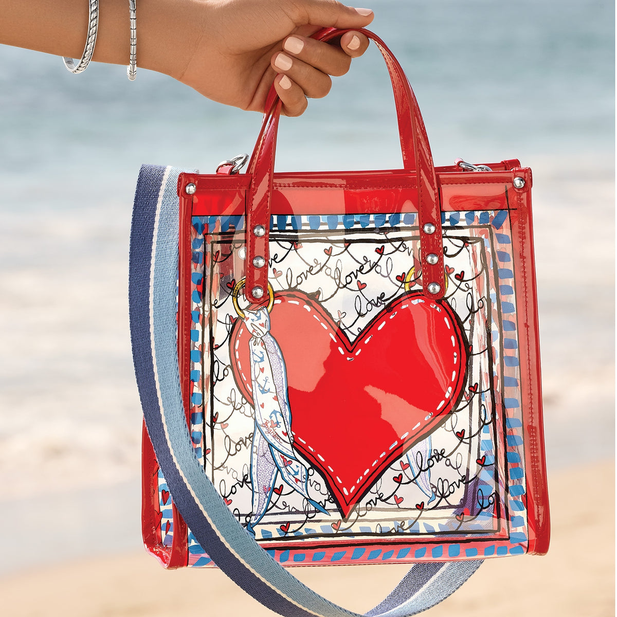 Brighton clear vinyl tote with a large red heart, 'love' script, and blue striped shoulder strap.