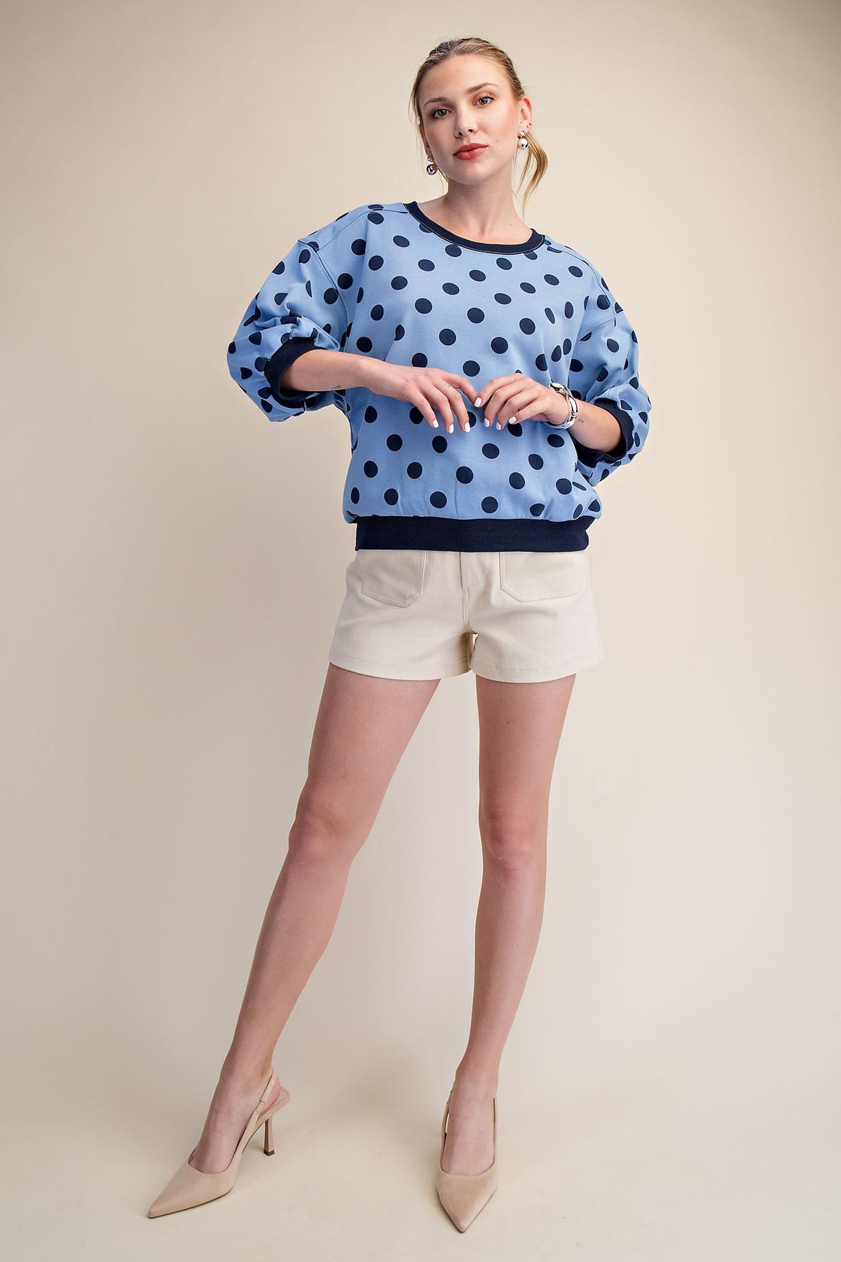 Woman wearing a light blue polka dot sweatshirt with bubble sleeves and cream shorts.