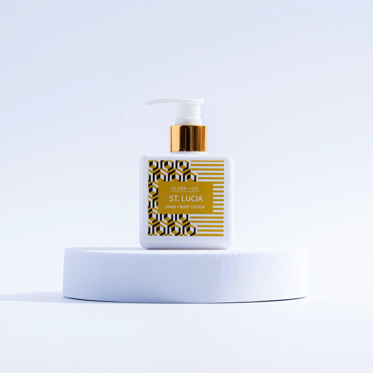 Oliver + Co. St. Lucia Hand + Body Lotion in a white square bottle with gold pump on a white podium.