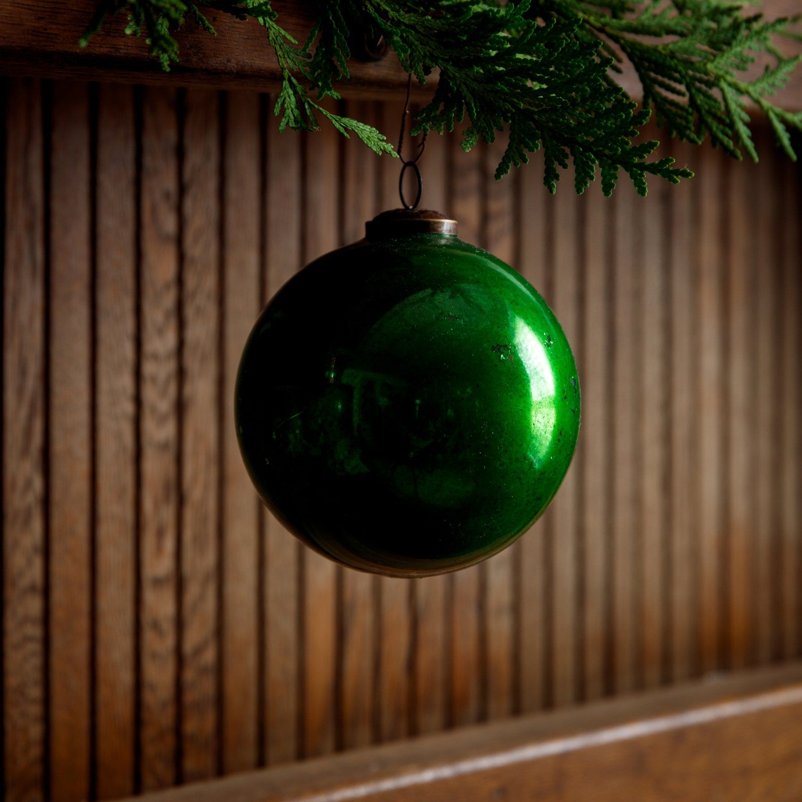 Vintage 5-inch green glass ball ornament hanging from a cedar branch against a wood panel wall.
