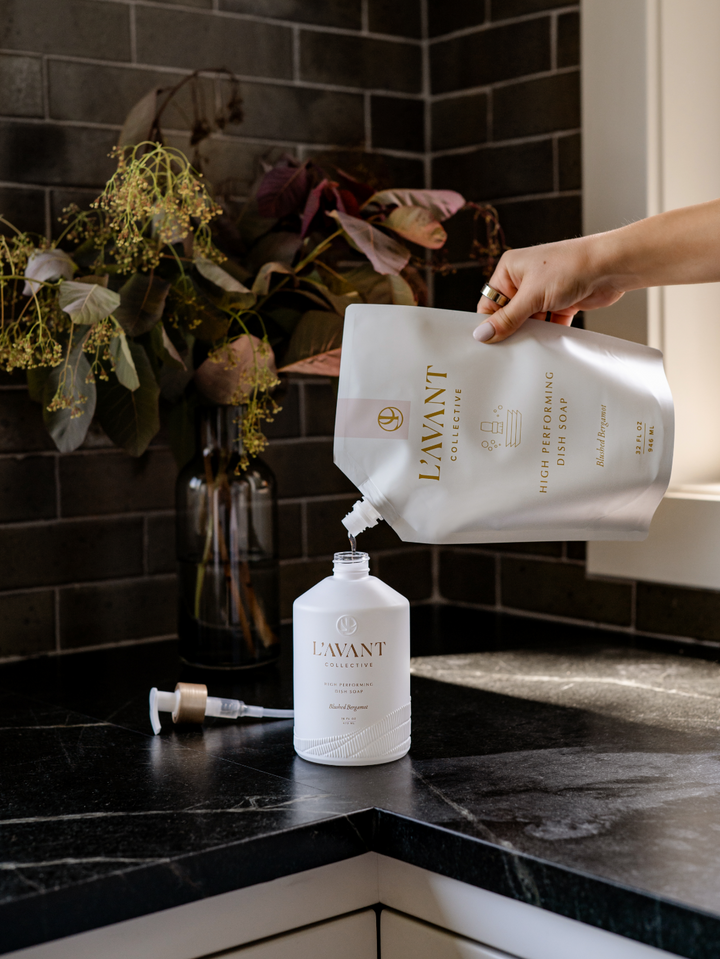 A hand pouring a 32 oz Blushed Bergamot dish soap refill pouch into a white L'AVANT glass bottle.