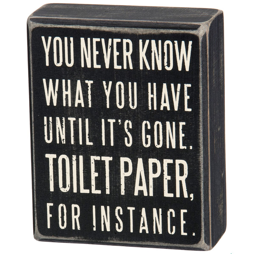 Black wooden box sign with white text reading 'You never know what you have until it's gone. Toilet paper, for instance.'