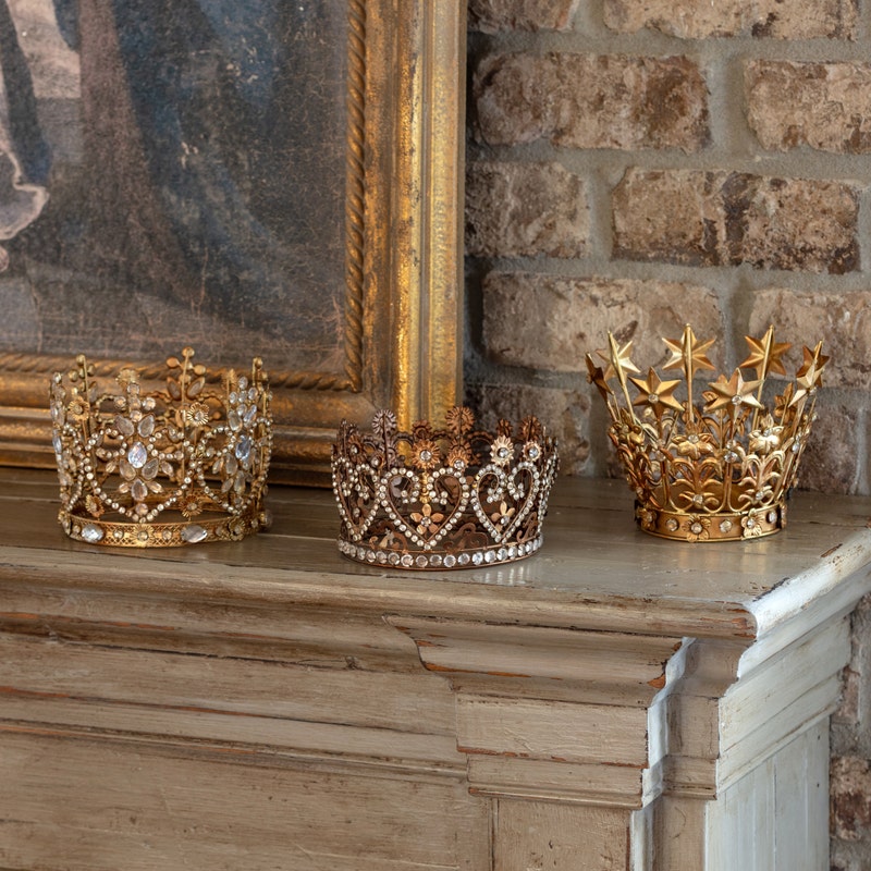 Three ornate, bejeweled gold and bronze King's Crowns displayed on a wooden mantel.