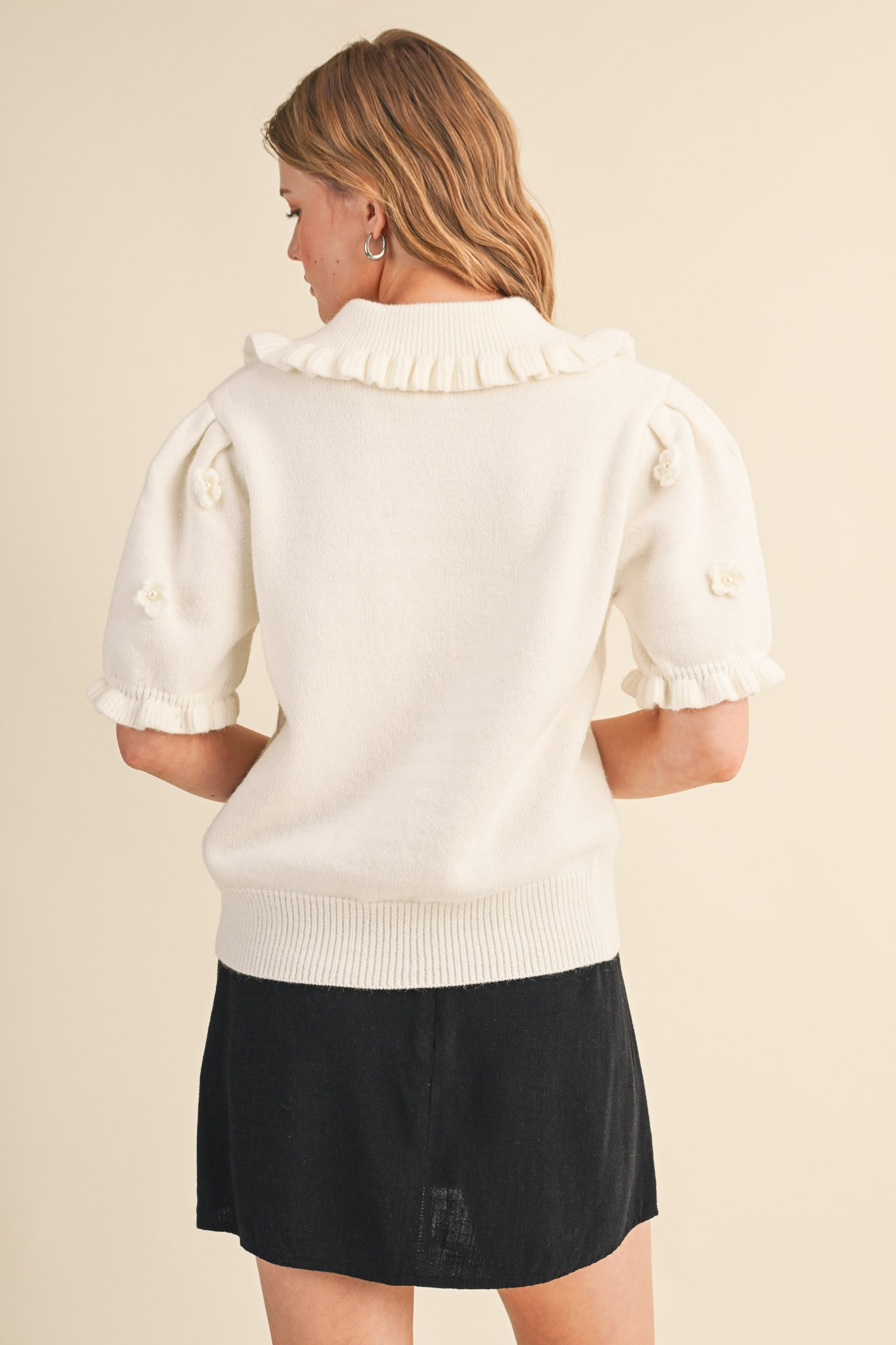 Back view of a white short-sleeved sweater with floral pearl appliqué and ruffled collar and cuffs.