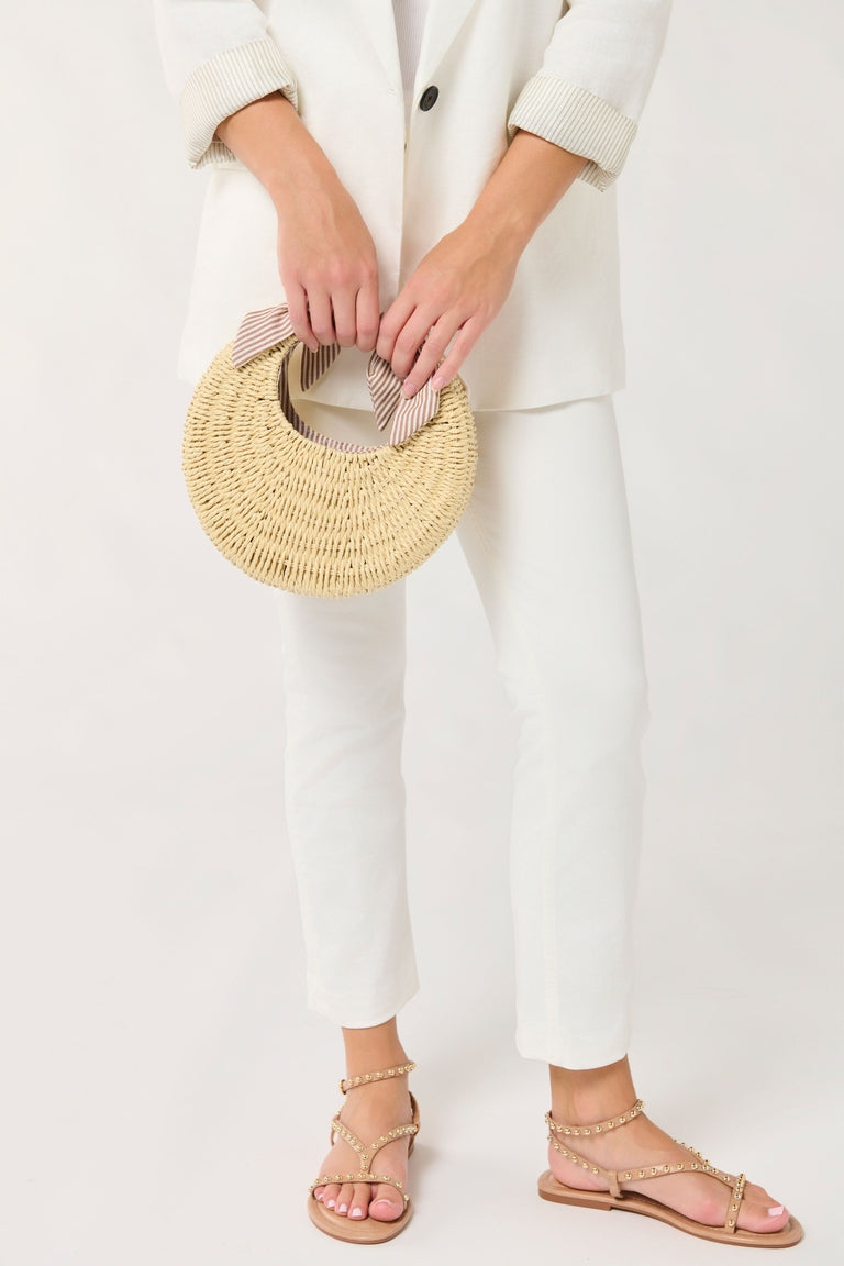 Straw Luna mini bag with striped ribbon detail, held by a person wearing white pants and sandals.