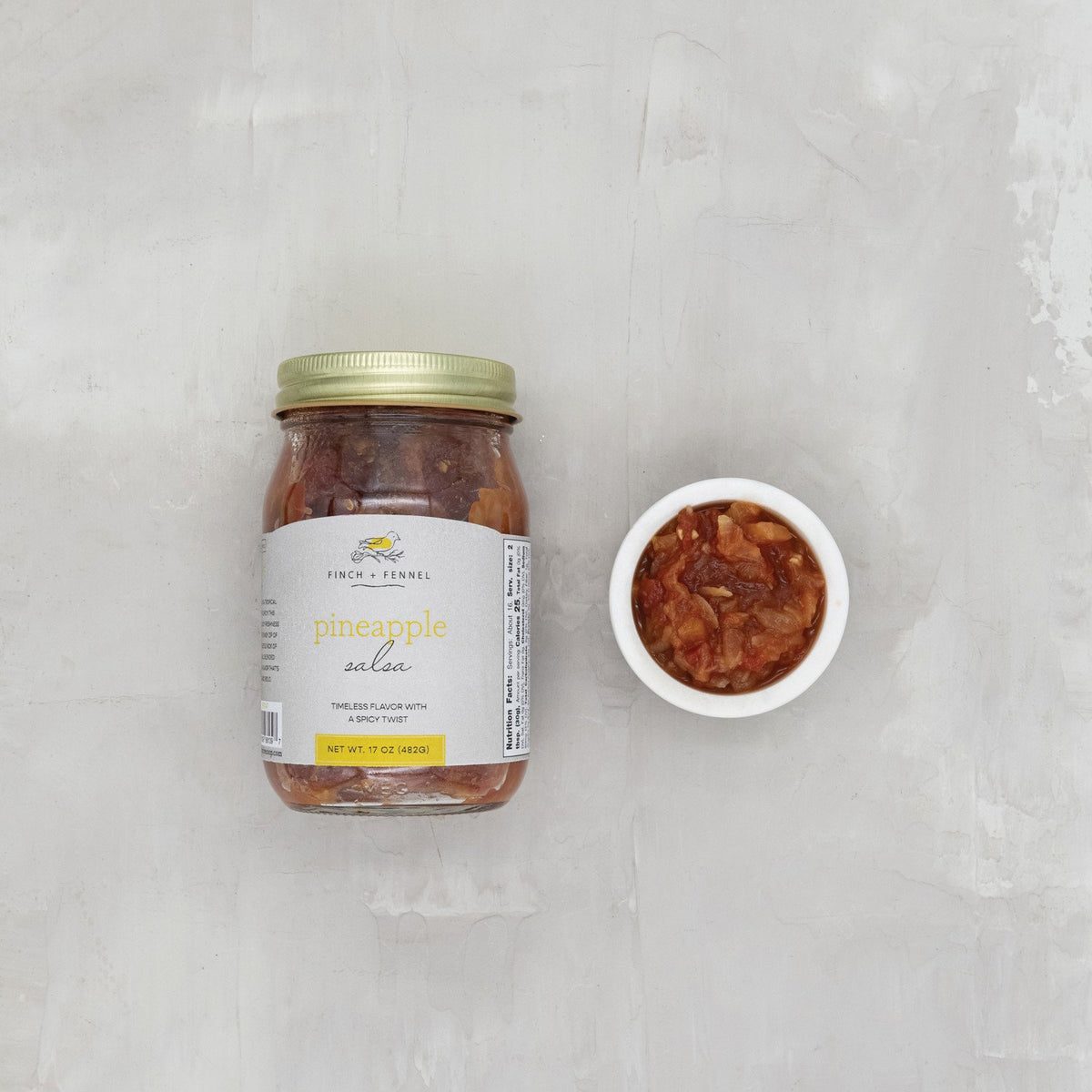 A 17 oz jar of Finch + Fennel Pineapple Salsa next to a small white bowl filled with the salsa.