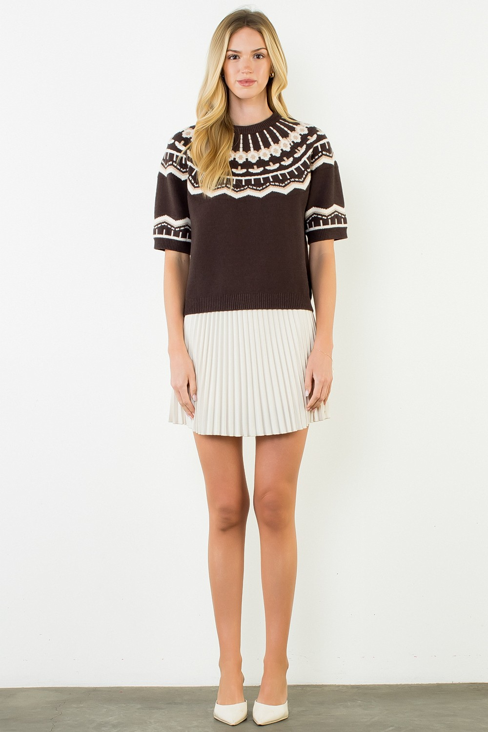 Woman wearing a brown short-sleeve sweater with a white geometric pattern and a pleated white skirt.