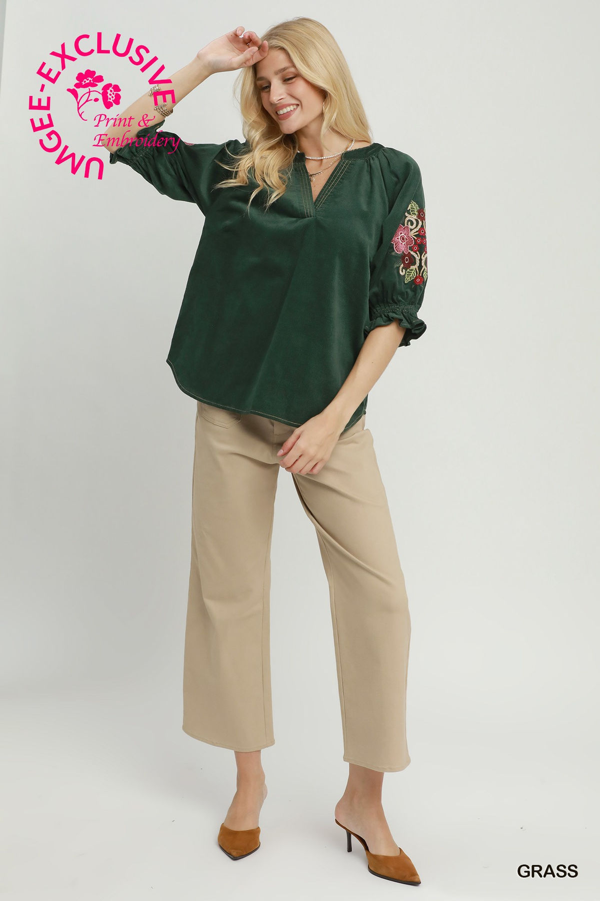 Woman wearing a dark green Eliana corduroy blouse with embroidered floral sleeves and tan trousers.
