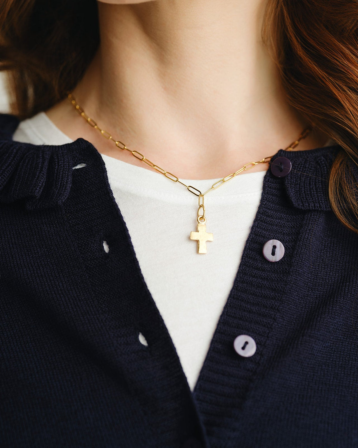 A person wearing a small gold cross pendant on a delicate paperclip chain necklace.
