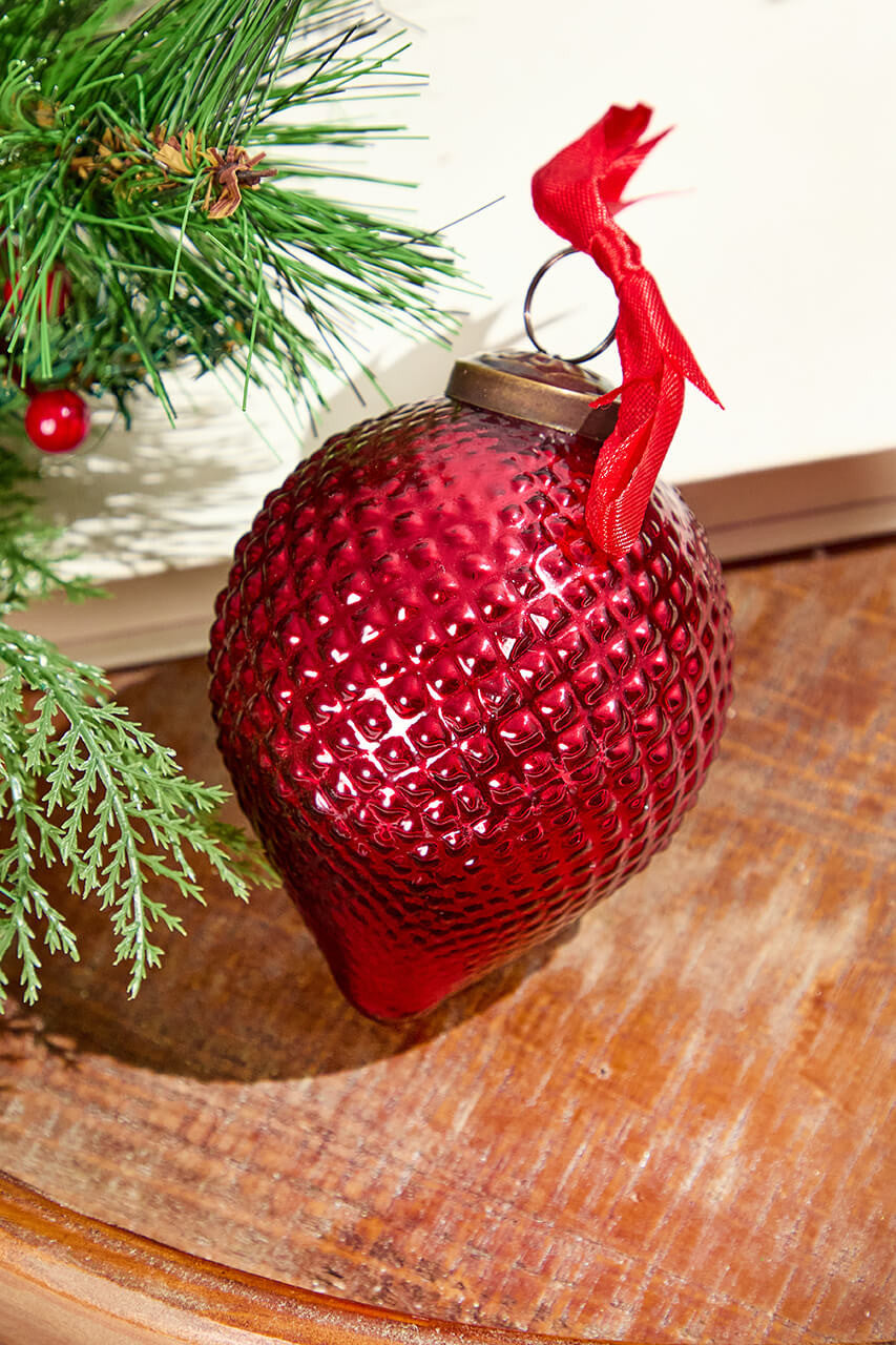 Cone Hobnail Ruby Glass 4" Ornament with a red ribbon on a wooden surface near pine branches.