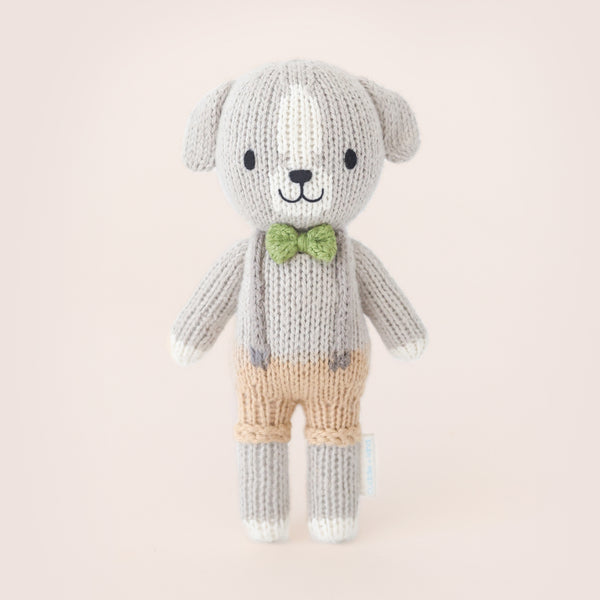 Hand-knit 6-inch grey dog doll wearing tan shorts, suspenders, and a green bow tie.