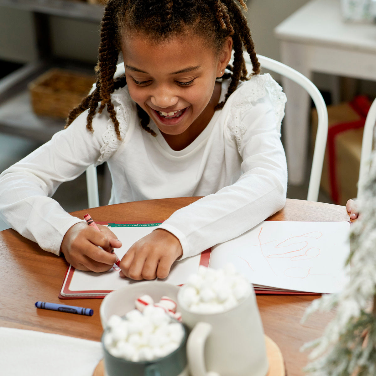 A young girl smiling while writing in the Santa's Kindness Journal with a red crayon.