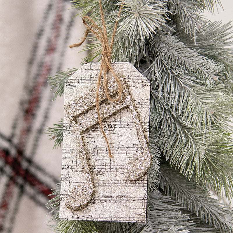 Glittered music note tag ornament with sheet music print and a jute hanger on a frosted pine branch.