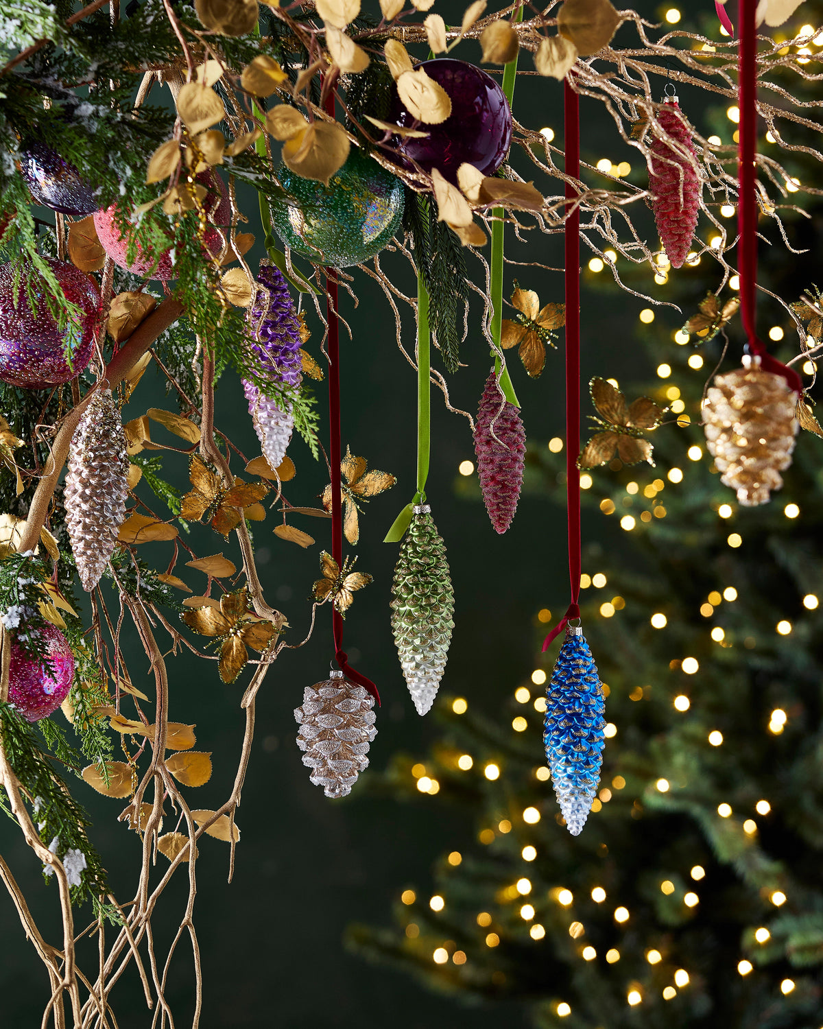Set of colorful glass pinecone ornaments in blue, green, and gold hanging from festive branches.