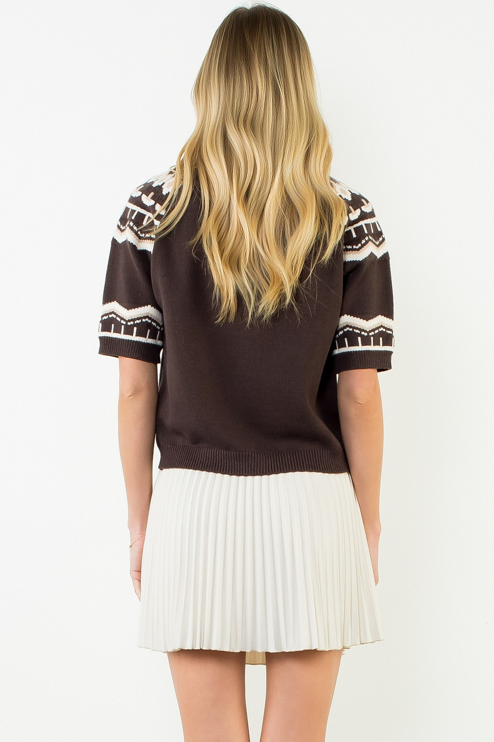 Back view of a woman wearing a brown short sleeve sweater with white patterns and a pleated skirt.