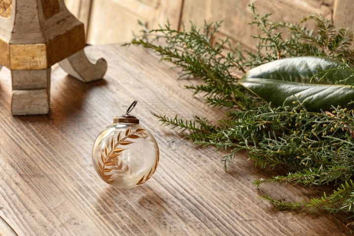 Golden Etched Laurel Glass 3" Ornament on a rustic wooden surface next to evergreen branches.