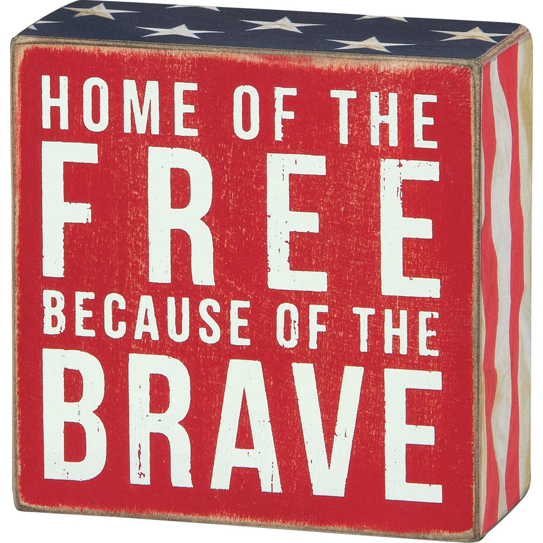 Red wooden box sign with white text "HOME OF THE FREE BECAUSE OF THE BRAVE" and patriotic stars and stripes.