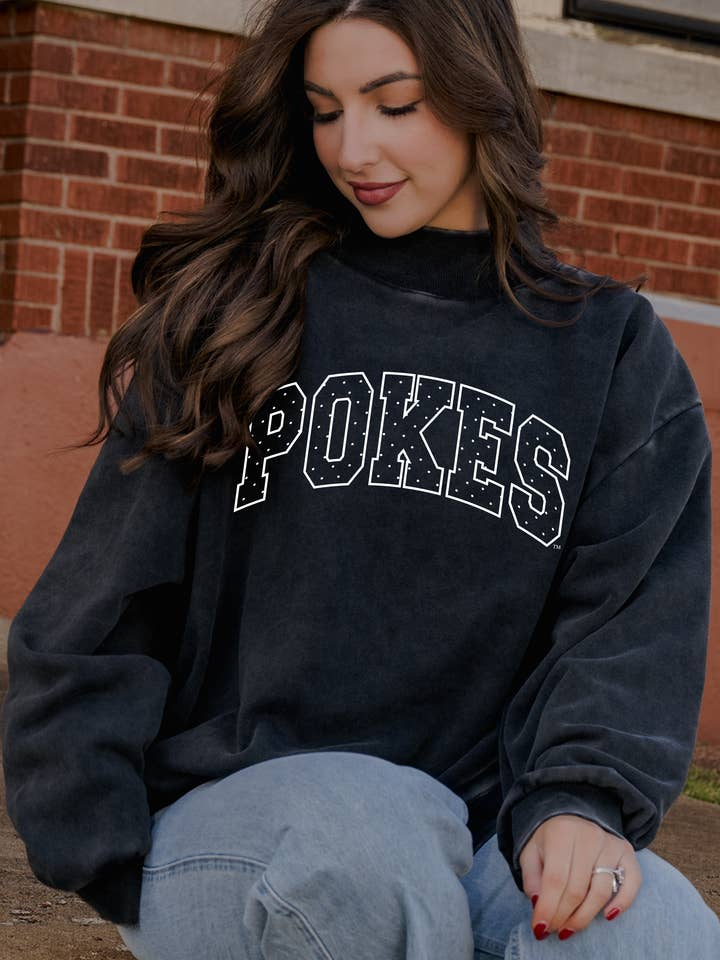 Woman wearing a charcoal grey mock neck sweatshirt with 'POKES' in white polka dot puff lettering.