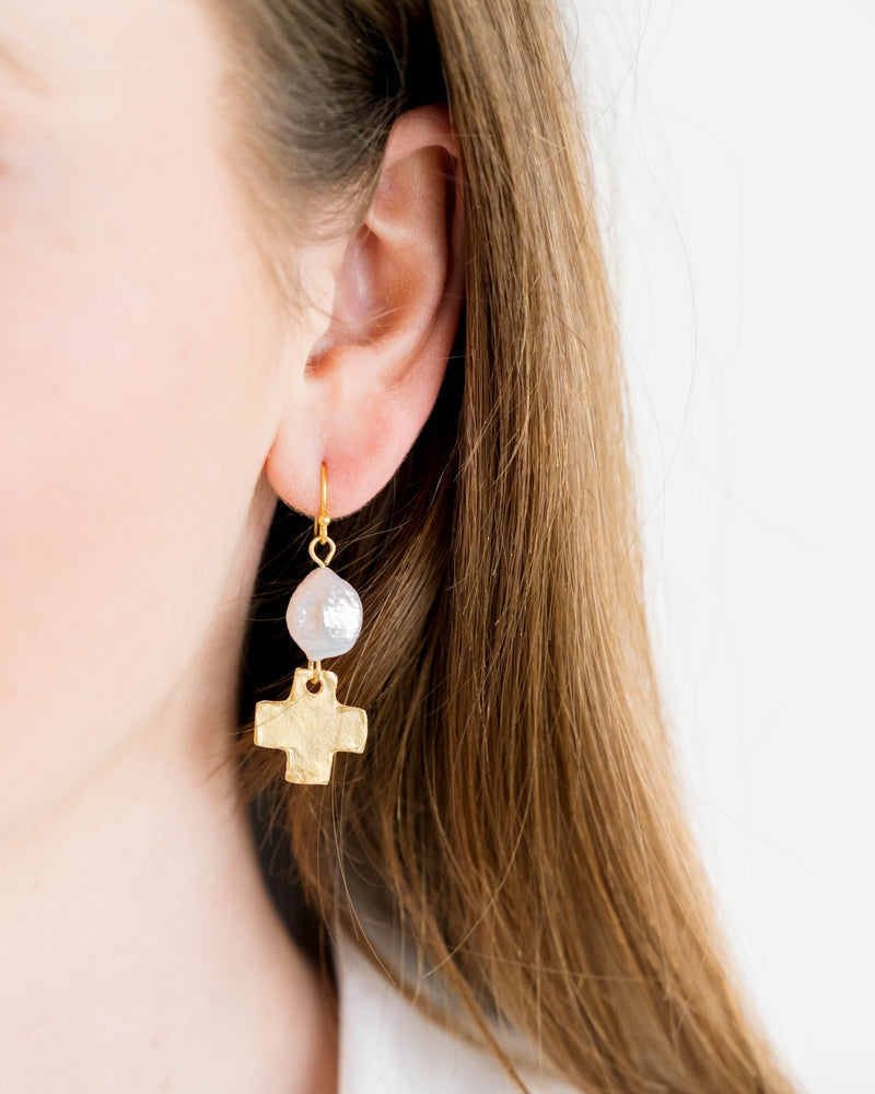 Close-up of a person wearing a gold fish hook earring with a white pearl and a gold cross charm.